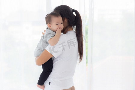年轻的亚洲母亲抱着小女婴在家里的卧室里一起散步，妈妈带着女儿和照顾，父母和孩子的关系，蹒跚学步的孩子和父母，室内，家庭观念。