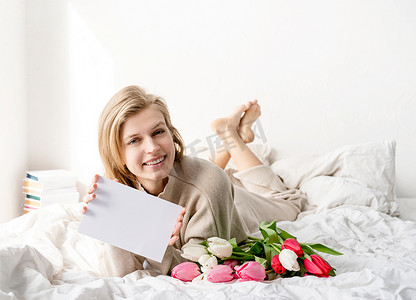 躺在床上的快乐女人穿着睡衣拿着郁金香花束和空白卡片