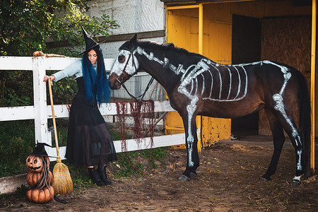 一个女巫摄影照片_一个女巫打扮的女孩站在畜栏的栅栏旁，旁边站着一匹画着骷髅的马