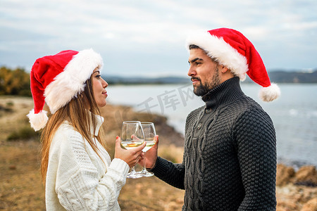美丽的异性恋情侣戴着圣诞帽，在户外举杯相望，手拿白葡萄酒杯，有选择地聚焦大海或海洋背景