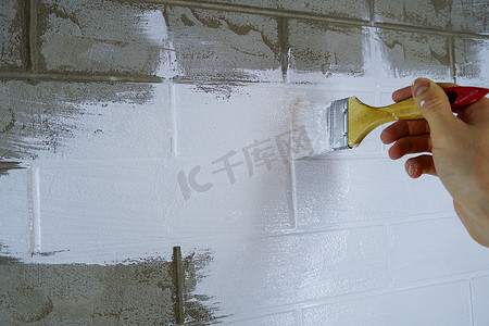 涂色画摄影照片_一名建筑工人在砖墙上涂漆。画家用刷子在墙壁上涂底漆，在房子或房间里进行维修。重建建筑，专家修复墙壁