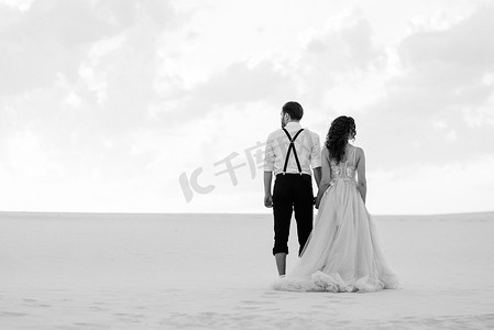 粉色沙滩天空摄影照片_一对年轻夫妇，一个穿黑色马裤的男人和一个穿粉红色连衣裙的女孩正沿着白色沙滩散步