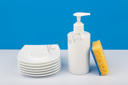 白色瓶中的洗碗洗涤剂、一堆碟子和蓝色背景中的清洁海绵