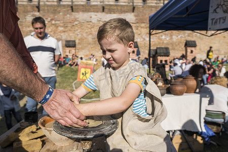 创意水壶摄影照片_乌克兰，卢茨克 — 2019 年 6 月 5 日：一个成年男子的手教孩子如何在美食节上用陶轮上的一块粘土制作一个水壶。