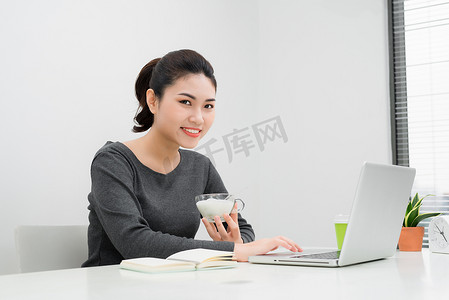 吃酸奶摄影照片_美丽的亚洲女商人坐在家里的工作地点，边吃酸奶边微笑