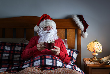圣圣诞老人摄影照片_圣诞老人在平安夜睡觉前坐在床上，端着一大杯热可可。