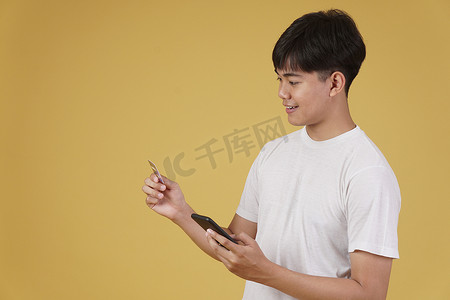 手持手机银行摄影照片_黄色背景中手持智能手机和信用卡在线购物的快乐亚洲青年肖像