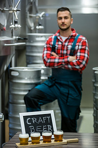 工厂标语牌摄影照片_啤酒取样器托盘上的四杯精酿啤酒和站在背景中的酿酒师