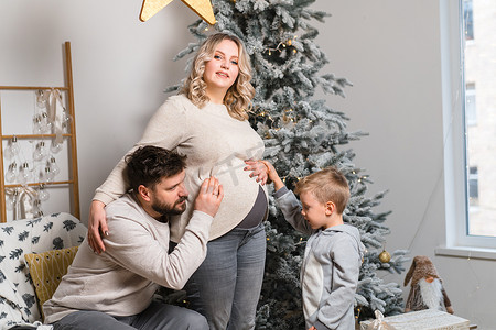 圣诞家庭幸福画像：爸爸、怀孕的妈妈和小儿子坐在家里的扶手椅上，靠近圣诞树拥抱微笑