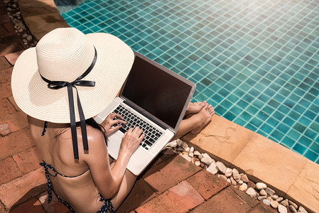 亚洲比基尼摄影照片_阳光明媚的日子里，亚洲年轻女性在泳池边使用带大帽子的笔记本电脑