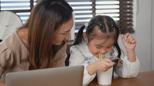 母亲和她的小女儿一起微笑着使用笔记本电脑在线学习和吃方便面