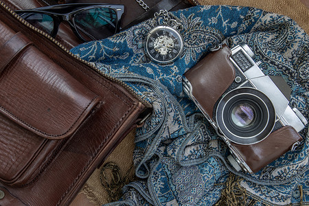 复古相机的顶部视图和带围巾、眼镜和怀表的棕色皮包在麻布背景上。