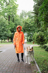 下雨狗摄影照片_穿着橙色雨衣的年轻女子和她的狗在公园里散步