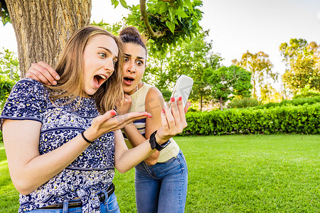 两个女孩最好的朋友惊讶地看着智能手机，脸上露出难以置信的表情，张大嘴巴，惊讶地睁大眼睛 年轻女性夫妇在城市公园享受社交网络的技术乐趣