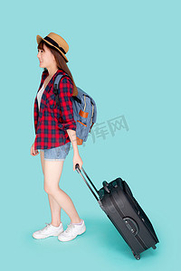 期中开课摄影照片_美丽的年轻亚洲女人拉着蓝色背景中突显的手提箱，有表情的亚洲女孩带着兴奋、旅行和旅行的概念快乐地拿着行李在假期中行走。