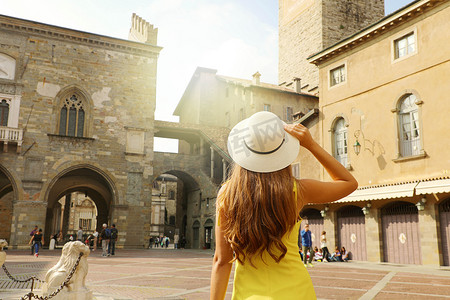 莫里尼摄影照片_戴帽子的漂亮女孩参观意大利贝加莫西塔阿尔塔中世纪古城的韦基亚广场广场。