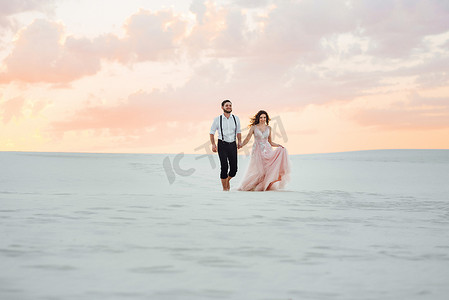 一对年轻夫妇，一个穿黑色马裤的男人和一个穿粉红色连衣裙的女孩正沿着白色沙滩散步