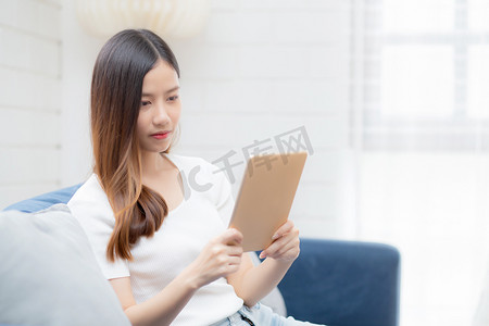 阅读交流摄影照片_年轻迷人的亚洲女性在家里的沙发上使用浏览平板电脑休息，快乐的女孩坐在沙发上放松阅读家里的数字小工具、交流和生活方式概念。