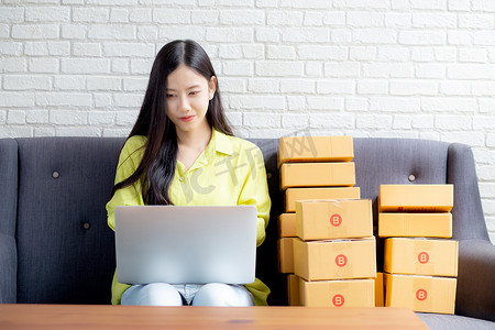 年轻的亚洲女性是商人，在沙发上使用笔记本电脑检查客户的订单，购买网上购物、商业中小企业或初创企业、企业家或卖家以及服务交付产品。