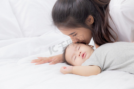 床上用品排版摄影照片_年轻的亚洲妈妈在卧室的床上亲吻小女孩的脸颊，妈妈爱新生儿和照顾，妈妈和孩子一起表达，父母和女儿，家庭观念。