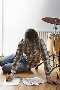 年轻人坐在架子鼓旁边的地板上写音乐