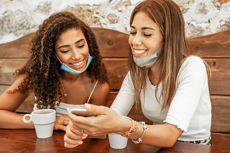 两个戴着防护面具的女孩坐在吧台旁，看着拿着咖啡杯的智能手机。