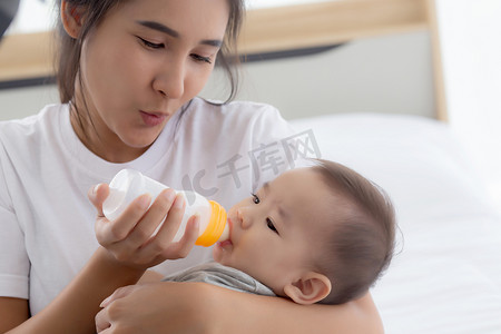 年轻的亚洲母亲在家里用奶瓶拥抱和喂养小女婴，新生儿的纯真与妈妈满意地喝酒，妈妈和孩子的关系和结合，家庭观念。