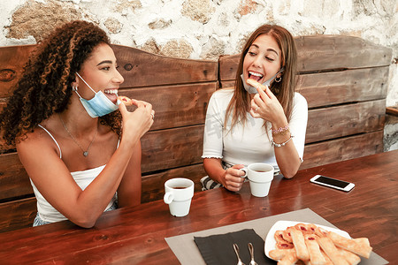 两位年轻漂亮的混血女性坐在户外质朴的木桌上，戴着低矮的防护面具，吃着一些糕点，拿着咖啡杯和智能手机面带微笑地看着对方