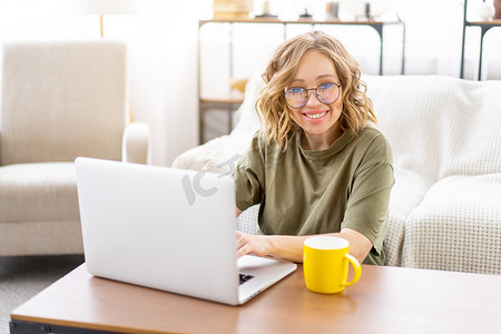 女性戴眼镜，坐在沙发旁，在家里的背景上使用笔记本电脑，沙发上有大窗户