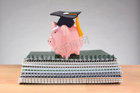 教师桌上一堆螺旋形笔记本的特写镜头，上面有一个戴着毕业帽的存钱罐，代表着高昂的教育成本。