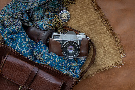 复古相机的顶部视图和带围巾、眼镜和怀表的棕色皮包在麻布背景上。