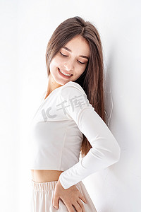 墙边摄影照片_穿着白色睡衣的快乐女孩站在家里的墙边微笑
