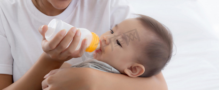 婴儿与奶瓶摄影照片_年轻的亚洲母亲在家里用奶瓶拥抱和喂养小女婴，新生儿的纯真与妈妈满意地喝酒，妈妈和孩子的关系和结合，家庭观念。