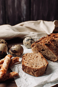 将深色无酵母荞麦面包放在羊皮纸上，旁边是鹌鹑蛋和意大利 grissini，放在木桌上的亚麻桌布上，采用乡村风格。