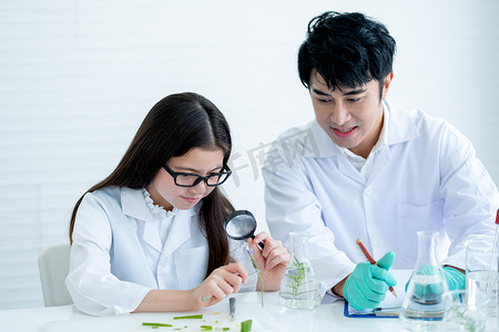 年轻的白人科学家女童透过一朵花，在实验室或教室的亚洲老师的指导和支持下，用放大镜看到更多细节。
