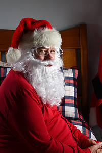 圣圣诞老人摄影照片_圣诞老人特写镜头坐在他的床上在他的红色长秋裤上，准备在圣诞节后睡觉。