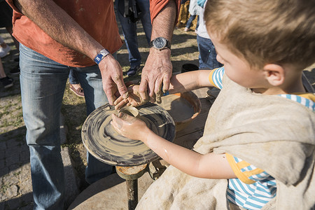 创意水壶摄影照片_乌克兰，卢茨克 — 2019 年 6 月 5 日：一个成年男子的手教孩子如何在美食节上用陶轮上的一块粘土制作一个水壶。