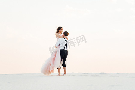 粉色沙滩摄影照片_一对年轻夫妇，一个穿黑色马裤的男人和一个穿粉红色连衣裙的女孩正沿着白色沙滩散步