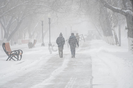 暴风雪国画摄影照片_一条积雪覆盖的道路，冬天在城市的恶劣天气下，人们在暴风雪、暴风雪或降雪中。北方的极端冬季天气条件。人们在大雪中穿过街道