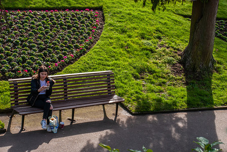 绿色唯美树叶摄影照片_在绿色公园的木凳上，一名年轻女子坐在手机上阅读短信。