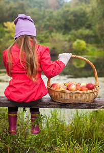坐在长凳上拿着大篮子和红苹果的小女孩的后视