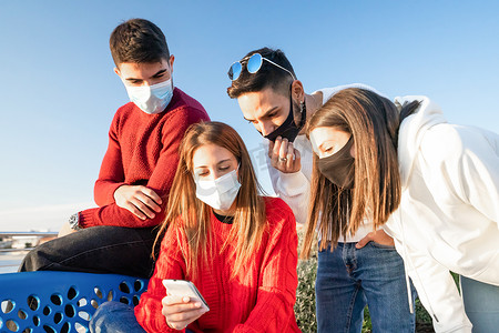 寒假安全摄影照片_一群戴着冠状病毒安全防护面具在户外看智能手机的年轻朋友 — 两对异性恋旅行者用手机在网上搜索他们的寒假