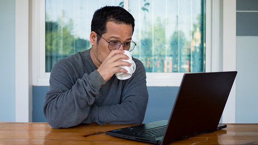 亚洲男人穿着长袖和眼镜。他正在喝咖啡，坐在家里的木桌上用电脑工作。