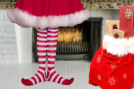 礼物长摄影照片_可爱可爱的小圣诞精灵女孩，脚尖，穿着条纹精灵长袜和红色连衣裙，站在燃烧的壁炉前，旁边是一个装满礼物的圣诞老人袋