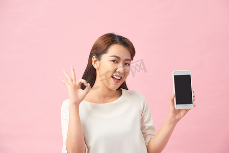 工作室拍摄摄影照片_快乐的亚洲肖像美丽可爱的年轻女性兴奋地拿着手机并打着 ok 的手势，工作室拍摄在粉红色背景中被隔离