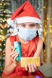 戴着医用面具、戴着圣诞帽和装饰背景的打开礼盒的女孩 — 礼物分享的概念、由于冠状病毒或 covid-19 大流行而导致的遥远的圣诞节庆祝活动。