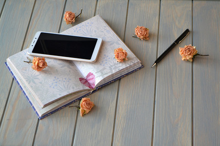 蓝灰色背景上的白色手机、笔、笔记本和复古玫瑰。