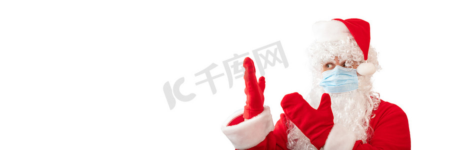 季中摄影照片_一个穿着圣诞老人服装、戴着医用面具、用双手指着他旁边的东西的男人的视图，在白色背景中被隔离。