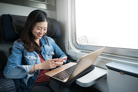 手机旅行摄影照片_亚洲女性在火车上拿着笔记本电脑对着智能手机微笑，在窗户上复制空间