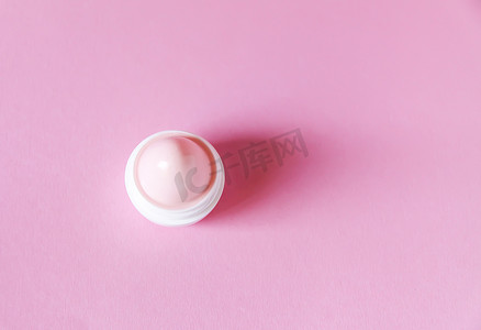 在柔和的粉红色背景上的白色小塑料容器中的润唇膏。